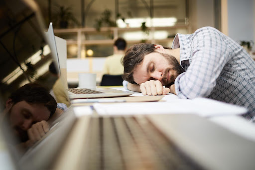 Man asleep at his desk beside a laptop in an office workspace.